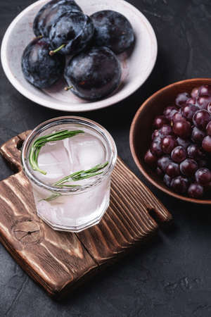 Fresh ice cold carbonated water in glass on cutting board with rosemary leaf near to wooden bowls with grape and plum fruits, dark stone background, angle viewの写真素材
