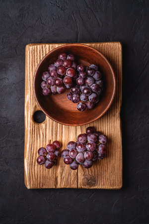 Fresh ripe grape berries in brown wooden bowl and cutting board on dark stone background, top viewの写真素材