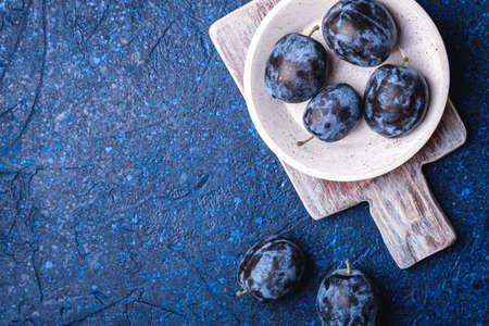 Fresh ripe plum fruits in white wooden bowl and old cutting board on blue abstract background, top view copy spaceの写真素材
