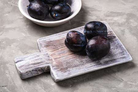 Fresh ripe plum fruits with water drops in wooden bowl and cutting board on stone concrete background, angle viewの写真素材