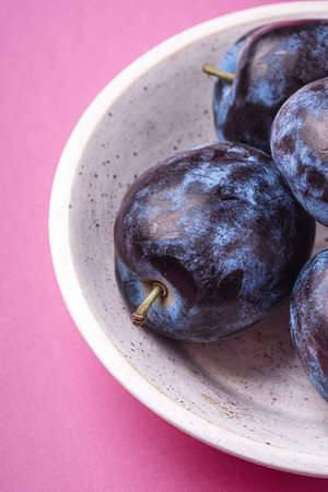 Fresh ripe plum fruits in white wooden bowl on pink minimal background, angle view macroの写真素材