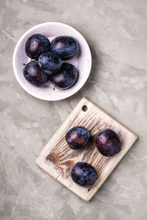 Fresh ripe plum fruits with water drops in wooden bowl and cutting board on stone concrete background, top viewの写真素材