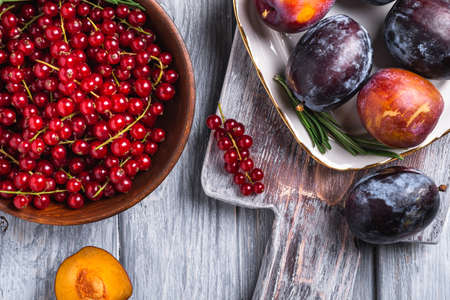 Fresh sweet plum fruits whole and sliced in plate with rosemary leaves on old cutting board with red currant berries in wooden bowl, grey wood background, top view close upの写真素材