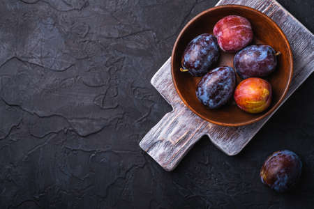 Fresh sweet plum fruits in brown wooden bowl on old cutting board, black textured background, top view copy spaceの写真素材