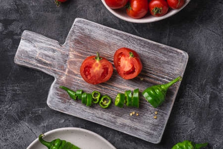 Fresh ripe tomatoes in bowl near to old wooden cutting board with hot chili peppers in plate, dark stone concrete background, top view close upの写真素材