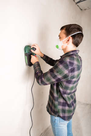 Man in protective mask working with electric sander to smooth plaster wall surface, room renovation conceptの写真素材