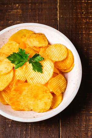 Fried corrugated golden potato chips with parlsey leaf in wooden bowl on wood backdrop, top viewの写真素材