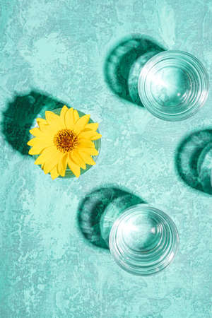 Fresh clear water drink with yellow flower in glass on turquoise background, hard light creative composition, top viewの写真素材