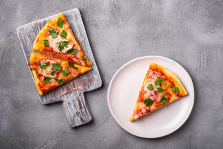 Hot pizza slices with mozzarella cheese, ham, tomato and parsley on wooden cutting board and plate, stone concrete background, top viewの写真素材