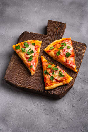 Hot pizza slices with mozzarella cheese, ham, tomato and parsley on brown wooden cutting board, stone concrete background, angle viewの写真素材