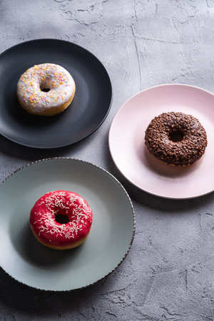 Three donuts on plates, chocolate, pink and vanilla donut with sprinkles, sweet glazed dessert food on concrete textured background, angle viewの写真素材