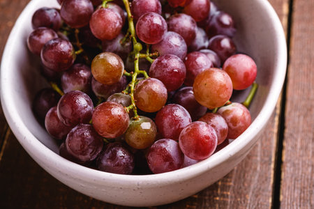 Fresh ripe grape berries in bowl on brown wooden background, angle view macro