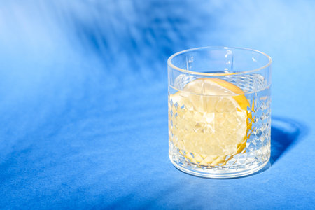 Glass with water and lemon on blue background with tropical palm leaf shadow