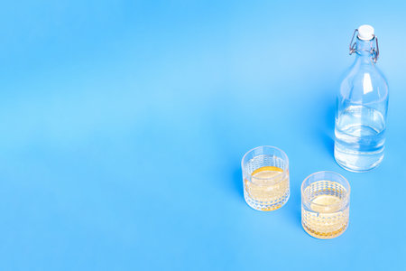 Glass with water and lemon near to bottle on blue background with copy space