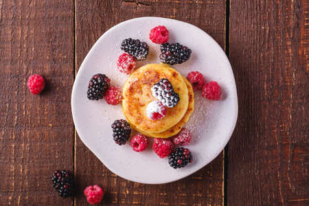 Cottage cheese pancakes and powdered sugar, curd fritters dessert with raspberry and blackberry berries in plate on dark brown wooden background, top viewの写真素材