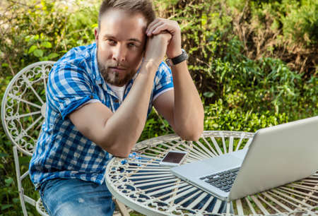 Young handsome man in casual clothes work at an iron table with computer against country garden.の写真素材
