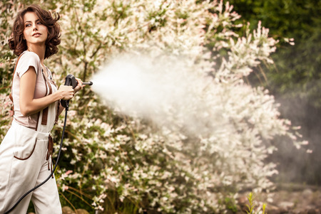 Outdoors portrait of beautiful  positive young woman in overalls which posing with water-cannon in summer garden.の写真素材
