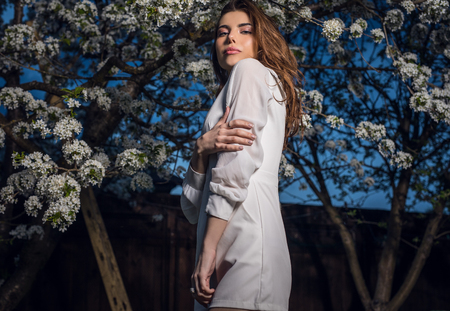 Portrait of beautiful young woman in white dress posing in autumn garden against blooming cherry.の写真素材