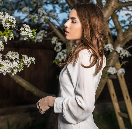 Portrait of beautiful young woman in white dress posing in autumn garden against blooming cherry.の写真素材