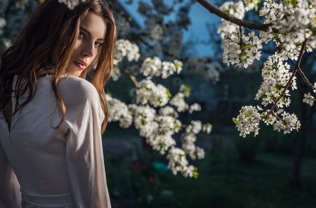 Portrait of beautiful young woman in white dress posing in autumn garden against blooming cherry.の写真素材