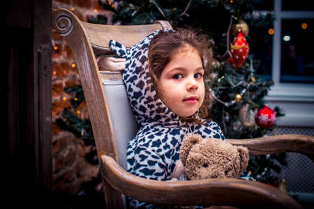 Pretty little girl smiling with teddy bear near the Christmas tree sitting in vintage chairの写真素材