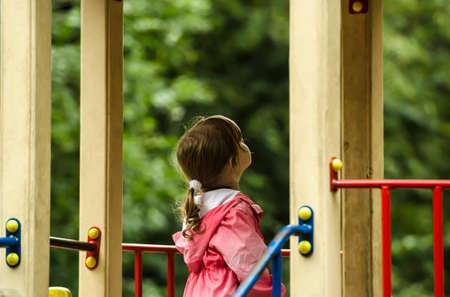 little girl looking at the sun in a pink jacket with a hood on the playgroundの写真素材