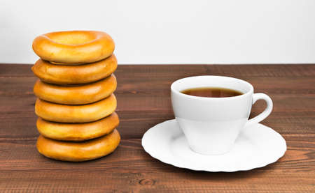 bunch of delicious rosy and bagels next to a white cup of tea on a saucer lie on a wooden tableの写真素材