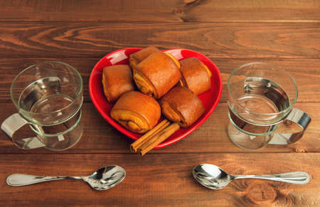 Two empty glass cups with metal handles and cinnamon rolls on a plate standing on a wooden tableの写真素材