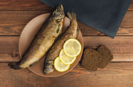 On the wooden table is a plate with two baked fish trout with lemon, two slices of rye bread and a napkinの写真素材