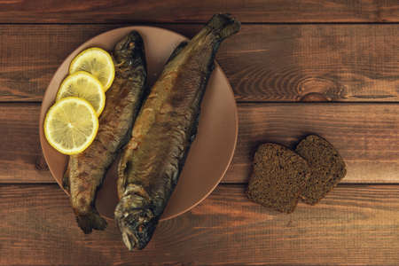On the wooden table is a plate with two baked fish trout with lemon, two slices of rye bread and a napkinの写真素材