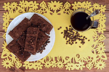 On a wooden table on yellow napkin glass cup of coffee with a spoon and a plate of chocolate truffle cake, a lot of coffee beansの写真素材