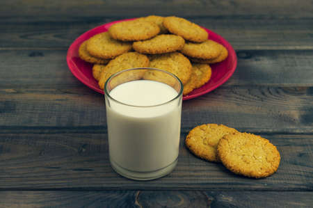On the rustic wooden table is breakfast - a red glass plate with homemade oat biscuits and a glass of milkの写真素材