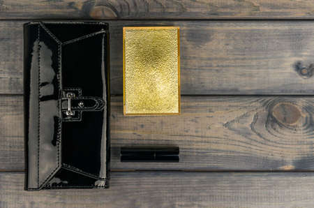 Purse-clutch black, powder box with mirror and lipstick golden lie on a wooden table background, there is a place for your text or imageの写真素材