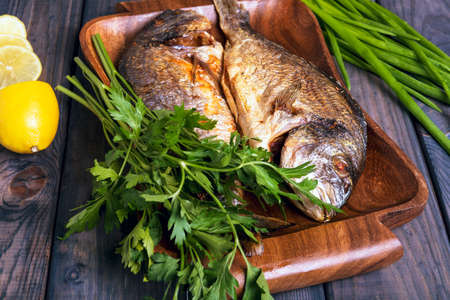 On a wooden table plate with two roasted carp fish dorado with parsley, chives and lemon.