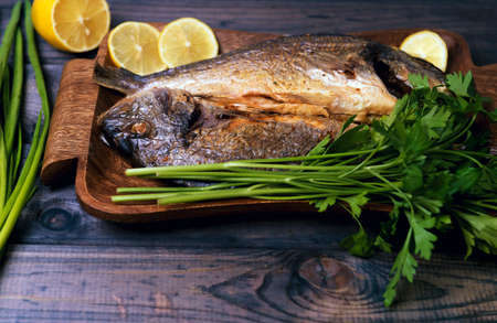 On a wooden table plate with two roasted carp fish dorado with parsley, chives and lemon.