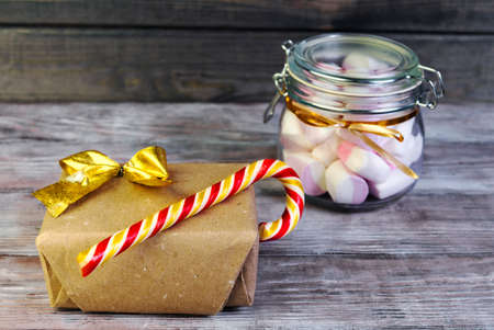 On a white shabby wooden table glass jar with a latch with a marshmallow and candy cane, Christmas gift wrapped in gold paper with ribbonの写真素材