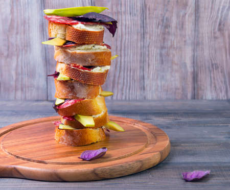 Small tapas sandwiches with bread, cheese, sausage meat, avocado and basil on a light wooden cutting board round table stacked pile in one large highの写真素材