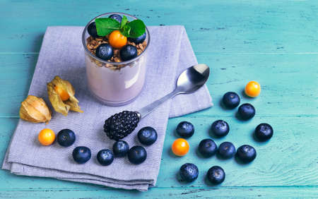 On a wooden blue background breakfast glass of yogurt, muesli, mint, blueberries and berries physalis, cotton clothの写真素材