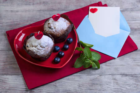 On wooden table in a bright red cloth with plate in the form of heart with two berry muffins with candles, sugar, blueberry, mint, envelope and sheet of paper, card Happy Valentine's Day, top viewの写真素材