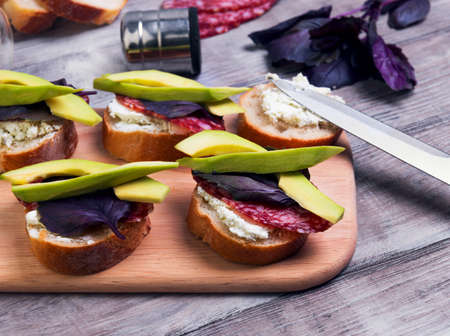 On a light wooden table cooking small sandwiches tapas with avocado, cheese, smoked sausage, basil, breadの写真素材