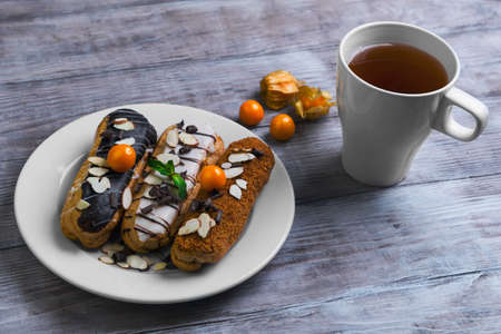 On a light wooden background in rustic style white round plate with cakes eclairs decorated with white and dark glaze, chocolate chips, mint, petals of almonds, a cup of tea, berries Physalisの写真素材