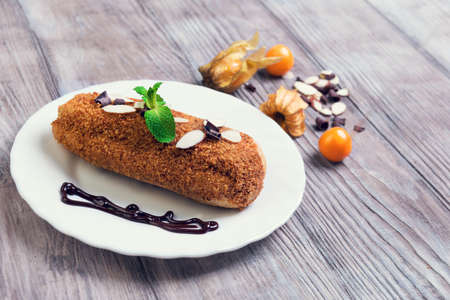 On a light wooden background white small plate with a cake decorated with chocolate eclairs, almonds, berries Physalisの写真素材