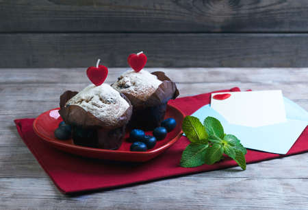 On wooden table in a bright red cloth with plate in the form of heart with two berry muffins with candles, sugar, blueberry, mint, envelope and sheet of paper, card Happy Valentine's Day, top viewの写真素材