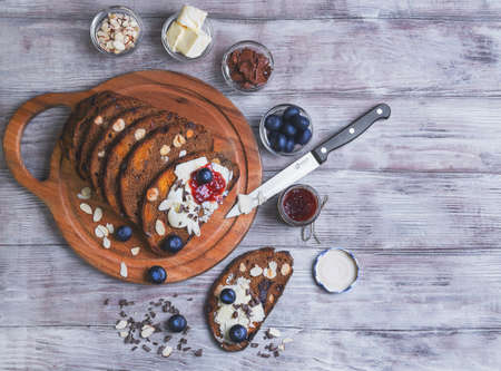 On a light wooden table sliced bread with fruits and nuts, blueberries, knife, chocolate paste, flakes, oil, almond petals, raspberry jam in a glass jar and a plate of rustic vintage styleの写真素材
