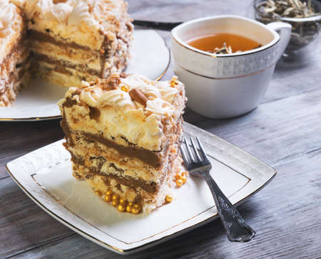 A large round cake of meringue and cocoa oil cream on a white plate, leaf tea, square cup, a piece of cake on a light wooden backgroundの写真素材