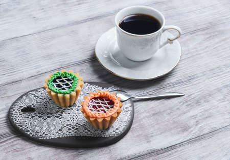Small cakes petit fours tartlets with jam and cream, ceramic board with a pattern of white lace, with a cup of coffee espresso on a light wooden backgroundの写真素材