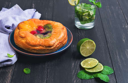 Denish fluffy bun with fruits and jelly on top, a glass of mojito, sliced lime, towel napkin, plate handmade ceramic on a dark wooden background in rustic styleの写真素材