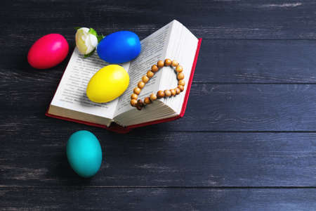Easter card, a small prayer bible, a rosary with a cross, painted eggs red, blue, green, yellow, white rose on a black wooden backgroundの写真素材