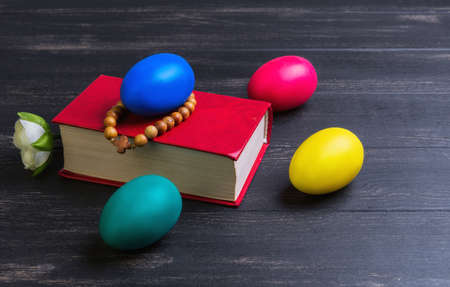 Easter card, a small prayer bible, a rosary with a cross, painted eggs red, blue, green, yellow, white rose on a black wooden backgroundの写真素材