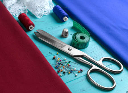 On a wooden table red and maroon cloth tape for removal of measures, tailor scissors, a metal thimble spool of thread with a needleの写真素材